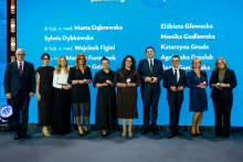 A group of people in formal attire is standing on stage in front of a blue backdrop with the inscription “Honored by the Minister of Health with the Honorary Badge ‘For Merits in Health Protection’” and a list of names. The participants are holding small red boxes, likely containing decorations.