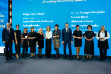A group of people in formal attire is standing on stage in front of a blue backdrop with the inscription “Honored by the Minister of Health with the Honorary Badge ‘For Merits in Health Protection’” and a list of names. The participants are holding small red boxes, likely containing decorations.