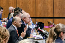 Conference participants are seated in rows in the lecture hall, with laptops, notebooks, and water bottles in front of them. One person is holding a microphone, taking part in the discussion.