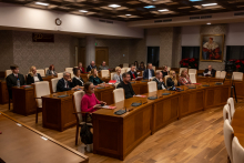 A conference room with wooden tables and light-colored chairs, where a group of people participating in the Nobel Session is seated; on the wall, a large portrait and decorative wallpaper are visible, while documents and microphones lie on the tables.