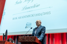 An elegantly dressed man is standing behind a lectern and speaking. In the background, there is a large multimedia screen displaying the text: “Crystal Lancet Gala. Academic Year 2024/2025.”