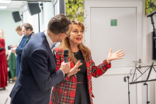 A man in an elegant suit is holding a microphone and talking to a woman dressed in a black dress and a red plaid jacket. The conversation is taking place in a bright room with white walls decorated with green leaves. In the background, a music stand and a microphone stand are visible.