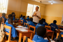 The teacher is standing in the classroom, holding a baby model in their hands, while the students in blue shirts are sitting at wooden desks with notebooks and books.