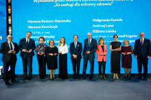 A group of people in formal attire is standing on stage in front of a blue backdrop with the inscription “Honored by the Minister of Health with the Honorary Badge ‘For Merits in Health Protection’” and a list of names. The participants are holding small red boxes, likely containing decorations.