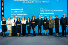 A group of people in formal attire is standing on stage in front of a blue backdrop with the inscription “Honored by the Minister of Health with the Honorary Badge ‘For Merits in Health Protection’” and a list of names. The participants are holding small red boxes, likely containing decorations.