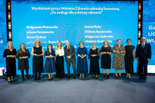 A group of people in formal attire is standing on stage in front of a blue backdrop with the inscription “Honored by the Minister of Health with the Honorary Badge ‘For Merits in Health Protection’” and a list of names. The participants are holding small red boxes, likely containing decorations.