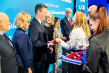 A group of people in formal attire is on stage during the UCK gala. In the foreground, a woman in a white shirt is handing a red case to a man in a suit.