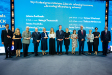 A group of people in formal attire is standing on stage in front of a blue backdrop with the inscription “Honored by the Minister of Health with the Honorary Badge ‘For Merits in Health Protection’” and a list of names. The participants are holding small red boxes, likely containing decorations.