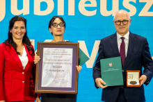Three people are standing in front of a blue backdrop with the word “Jubilee.” The person on the left is wearing a red outfit, the one in the middle is holding a framed diploma, and the person on the right has a green folder and an open box with a medal.