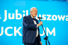 A person in a dark suit with a bow tie is standing at a microphone and speaking. In the background, there is a light blue screen with large white lettering.