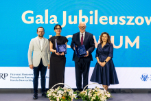 Four people are standing on stage in front of a large screen with the inscription “Jubilee Gala.” Two of them are holding plaques or awards. At the bottom of the stage, floral decorations are visible.