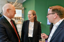 Three people are standing and talking in a modern interior with a green wall. One person is holding a cup with a spoon and has a lanyard with an ID badge hanging around their neck.