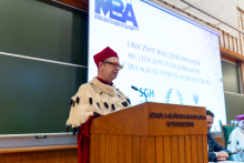 A person dressed in a rector's robe with a red cap and a decorative chain stands at a wooden lectern. In the background, there is a large screen with the inscription “Ceremonial presentation of diplomas to participants of the 6th edition of the Executive SGH WUM MBA in Healthcare program.” 