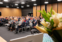 A conference hall filled with participants seated in rows on chairs, dressed in elegant business attire. In the background, wooden panels and doors are visible, with people standing nearby observing the event. In the foreground, there is a bouquet of white flowers in a vase, partially obscuring the view of the hall.