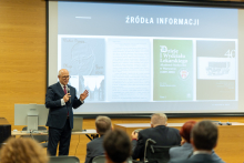 Professor Rafał Krenke is speaking and presenting slides on a large multimedia screen.