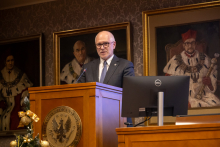 A person in a suit stands behind a wooden lectern bearing the emblem of the medical university, delivering a speech in a hall adorned with portraits of rectors in historical attire. A microphone is placed on the lectern, and a monitor is positioned nearby. In the background, elegant picture frames are visible, while festive decorations appear in the foreground.