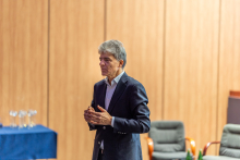 An elegantly dressed man is standing in the center of an auditorium and speaking. In the distance, there is a table with crystal statuettes on it.
