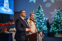 A smartly dressed young woman, Anna Kupczak, and a young man, Karol Sadowski, are standing at a lectern. The man is speaking into a microphone. In the background, three Christmas trees decorated with white baubles and lights are shining.