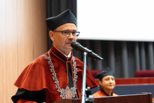 The dean of the faculty, dressed in a black and orange gown, is standing at the lectern and speaking.