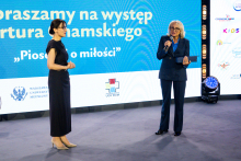 Two people are standing on stage in front of a large screen with the inscription “Join us for Artur Chamski’s performance ‘A Song About Love.’” One person is holding a microphone and speaking, while the other stands with their hands clasped.