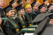 People are sitting in a wooden auditorium during a ceremony, dressed in black gowns with green trim and black caps with tassels. In the center, there is a person wearing a yellow cap, and the gowns bear the inscription “Edition VI” and the MBA program logo. 