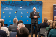 A man, the Rector of WUM, standing on stage in a conference hall, dressed in a dark suit, with his hands clasped in front of him, positioned by a microphone on a stand. In the background, there is a blue backdrop featuring logos and inscriptions highlighting the anniversaries.