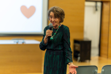 An elegantly dressed woman, Professor Ryszarda Chazan, is standing and speaking into a microphone. In the background, a multimedia screen is visible.