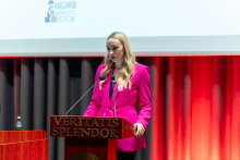 A young, elegantly dressed woman is standing behind a lectern and speaking. In the background, there is a multimedia screen.