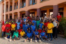 A large group of children in colorful shirts – red, yellow, green, and blue – are standing and sitting in front of a building with an orange facade and balconies, posing for a group photo with adults.