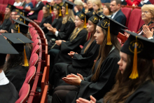 A group of graduates dressed in gowns is sitting on red seats in the auditorium, applauding.