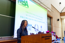 A person is standing at a wooden lectern. In the background, there is a large screen with the inscription “Ceremonial presentation of diplomas to participants of the 6th edition of the Executive SGH WUM MBA program in Healthcare.” 