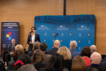 A woman, the Director of UCK, standing on stage in a conference hall, dressed in an elegant outfit, holding a microphone in her hands. Next to her, there is another microphone on a stand. In the background, a blue backdrop is visible with inscriptions highlighting the anniversaries.