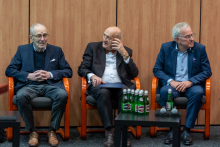 Three elegantly dressed men are sitting in armchairs, with small tables in front of them holding bottles of water.