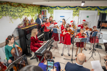 A group of children in festive outfits is performing a concert in a small hall, playing pieces on violins and cellos accompanied by a piano. In the background, there is a decoration made of green leaves and garlands, and Christmas figurines are placed on the piano.