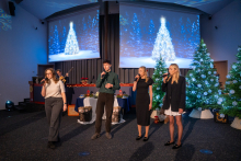 Four people are standing on stage and singing into microphones. In the background, there are two Christmas trees decorated with white baubles and lights, as well as two large screens displaying an image of a bright Christmas tree in a winter forest. On the left side, a person is playing the guitar. The hall is decorated with festive elements.