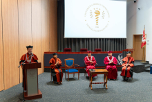 The stage of the auditorium: behind the lectern stands the dean, delivering a speech. Next to him are members of the faculty authorities and the vice-rector, all wearing academic gowns. In the background, there is a multimedia screen displaying the yellow logo of the Faculty of Pharmacy.