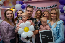 A group of people posing for a photo. In the background, behind them, you can see white and purple balloons and a ribbon with the words “Premature Baby Day.”