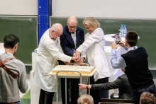 Three people – two wearing white lab coats and one in a suit – are jointly cutting a large rectangular cake placed on a metal cart.