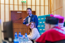 A man in a navy blue plaid jacket stands at a wooden lectern, holding a microphone. An elegantly dressed woman sits next to him at a table.