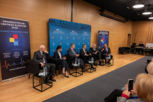 A discussion panel in a conference hall with six participants seated in black armchairs arranged in a row. Each armchair has a small table in front of it with water bottles and glasses. In the background, there is a blue backdrop featuring inscriptions highlighting the anniversaries.