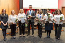 Seven people are posing for a photo. They are all elegantly dressed. Five of them are holding a single flower and a diploma, while two additionally have crystal statuettes.