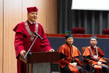 The vice-rector, dressed in a red gown and cap, is speaking into the microphone from behind the lectern. Next to him are seated the dean and the vice-dean.