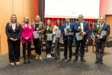 Eight people are posing for a photo. They are all elegantly dressed. Five of them are holding a single flower and a diploma, while two additionally have crystal statuettes.