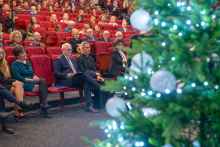 Several rows of people are seated in the hall on red chairs, looking toward the stage. In the foreground, part of a Christmas tree decorated with white baubles and lights is visible. Dozens of people are present in the hall, participating in the event.