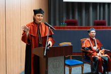 Vice-Dean Professor Olga Ciepiela is speaking into the microphone from behind the lectern. She is wearing a black and orange gown. Her right hand is raised. Next to her, the dean is seated.
