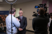 An interview in a hallway, where one person is holding a microphone with the logo of the television station “TVP” and talking to another person dressed in an elegant suit. Next to them, a camera operator is recording the conversation, and above them, a ring light is set up for illumination.