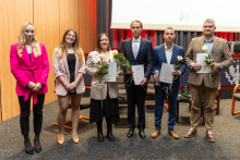 Six people are posing for a photo. They are all elegantly dressed. Five of them are holding a single flower and a diploma, while two additionally have crystal statuettes.