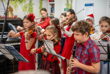 A group of children in red outfits is playing the violin and clarinet during a Christmas concert. In front of them are music stands, and in the background, a piano and green plant decorations can be seen, emphasizing the festive character of the event.