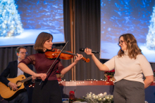 Two people are standing on stage. One is playing the violin, and the other is holding a microphone directed toward them. In the background, a person is playing the guitar, and two large screens display an image of a bright Christmas tree in a winter forest.