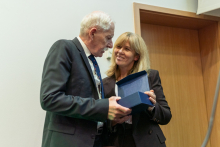 Dwie osoby, po lewej elegancko ubrany mężczyzna, prof. Rowiński, po lewej kobieta. Stoją blisko siebie. Trzymają w dłoniach granatowe pudełko z odznaczeniem dla prof. Rowińskiego. Patrzą na siebie.